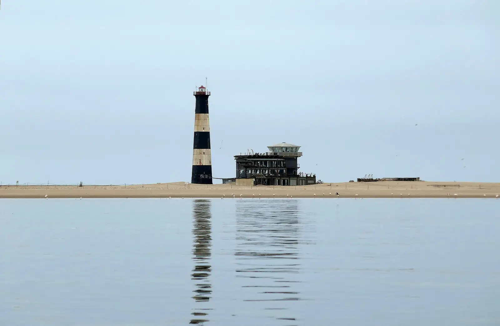 Black-and-white striped Pelican Point lighthouse and lodge on the sand spit, reflected in calm water